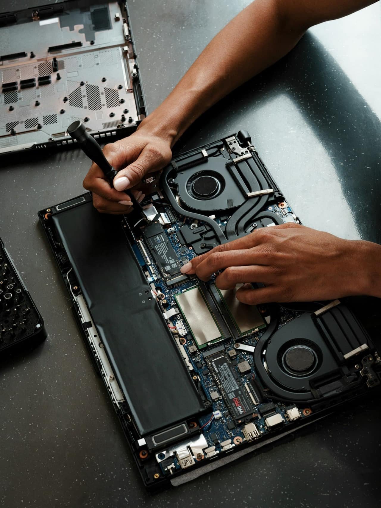 A technician opening a laptop's back panel with a screwdriver to reach the dusty cooling fan